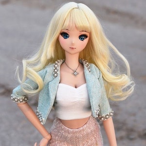 May include: A blonde doll with blue eyes wearing a light blue jacket with pearl accents, a white crop top, and a pink skirt.