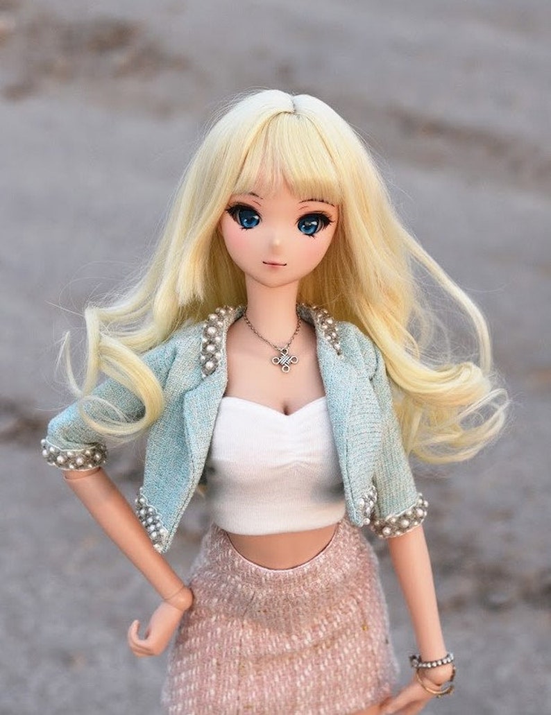 May include: A blonde doll with blue eyes wearing a light blue jacket with pearl accents, a white crop top, and a pink skirt.