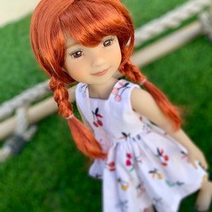 May include: A red-haired doll wearing a white dress with a cherry print. The doll has a sweet expression and is looking up at the camera.