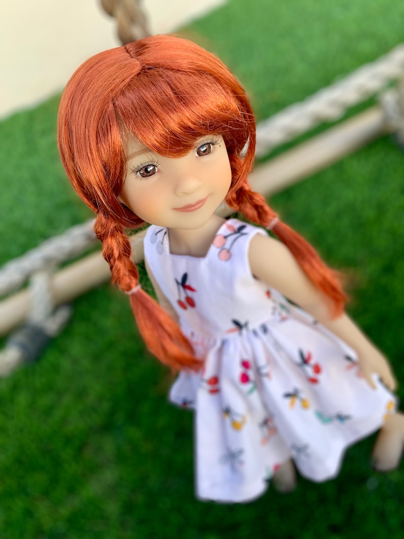 May include: A red-haired doll wearing a white dress with a cherry print. The doll has a sweet expression and is looking up at the camera.