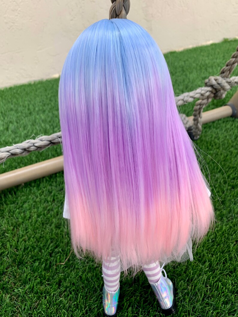 May include: A doll with long, straight, ombre hair that transitions from light blue to purple to pink. The doll is wearing a white dress with a pink tulle skirt and white and purple striped tights.