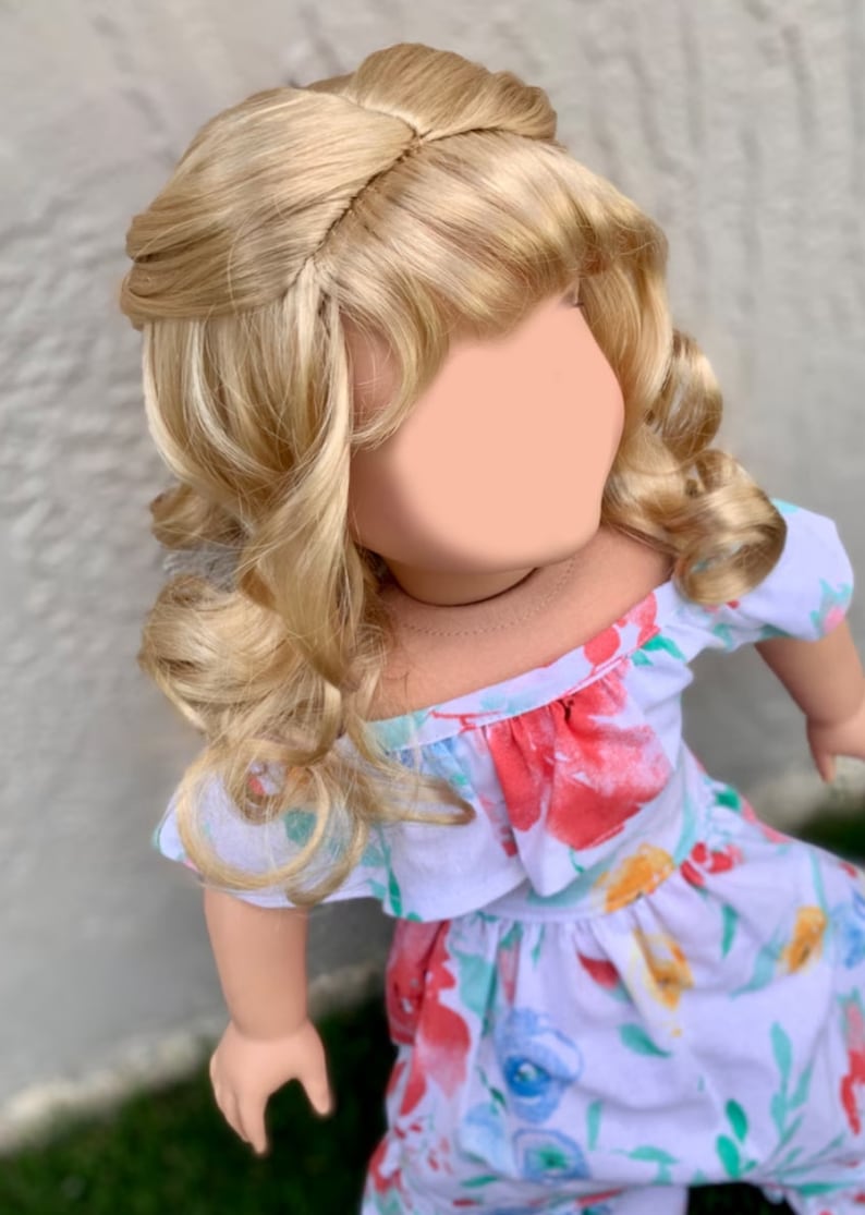 May include: A doll with blonde, wavy hair styled in an updo. The doll is wearing a white dress with a floral pattern in red, blue, and yellow. The dress has an off-the-shoulder neckline.