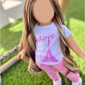 May include: A doll with long brown and blonde hair wearing a white t-shirt with a pink Eiffel Tower graphic and the word "adore" printed on it. The doll is also wearing pink pants and white sneakers.