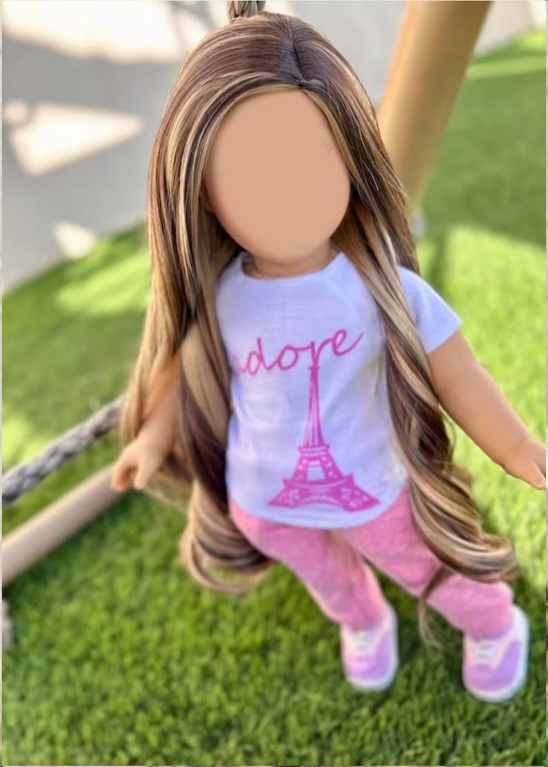 May include: A doll with long brown and blonde hair wearing a white t-shirt with a pink Eiffel Tower graphic and the word "adore" printed on it. The doll is also wearing pink pants and white sneakers.