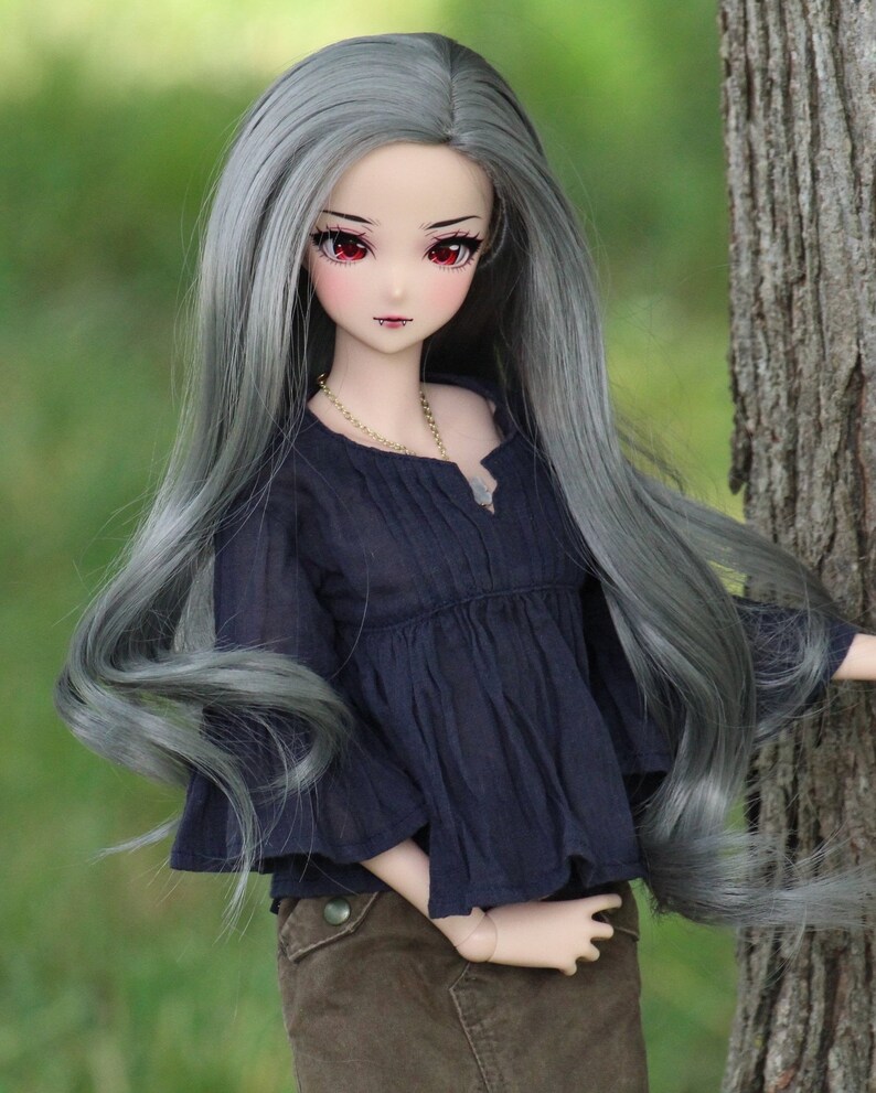 May include: A doll with long, flowing silver hair, wearing a dark blue blouse and brown pants. The doll has red eyes and a delicate face.
