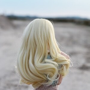 May include: A doll with long, flowing blonde hair, wearing a light pink dress. The doll is standing in a sandy area with a blurred background.