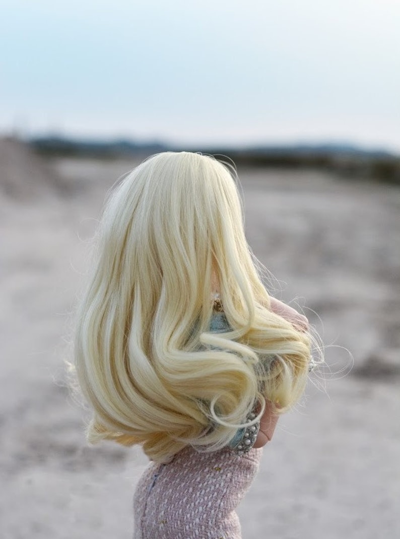 May include: A doll with long, flowing blonde hair, wearing a light pink dress. The doll is standing in a sandy area with a blurred background.