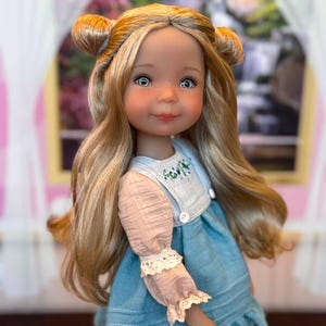 Op de afbeelding: Een pop met lang blond haar in twee knotjes, blauwe ogen en sproeten. De pop draagt een blauwe jurk met een wit lijfje, kleine witte knoopjes en lange, lichtroze mouwen met kanten randjes bij de boorden en de zoom van de jurk.