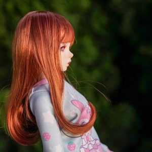May include: A doll with long, flowing red hair wearing a light blue and pink patterned dress. The doll is looking to the right and has a soft, feminine expression.