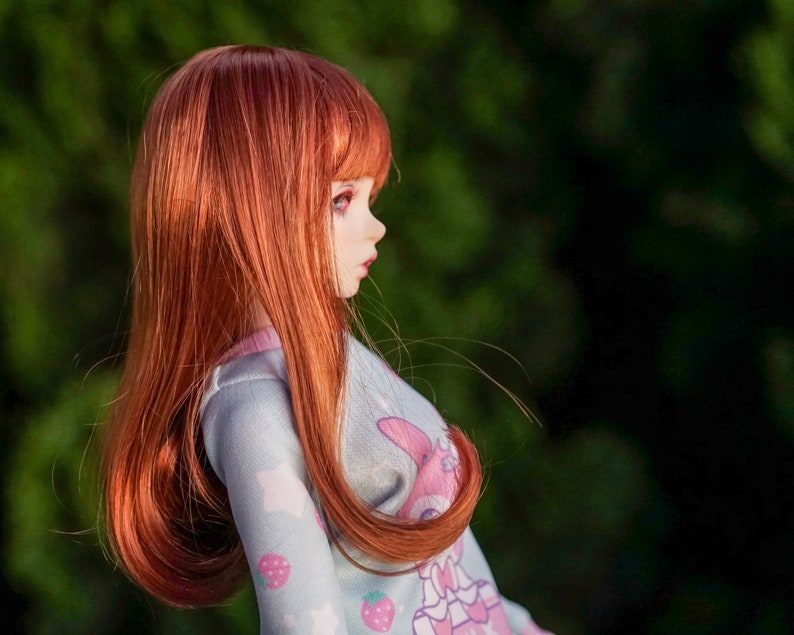 May include: A doll with long, flowing red hair wearing a light blue and pink patterned dress. The doll is looking to the right and has a soft, feminine expression.