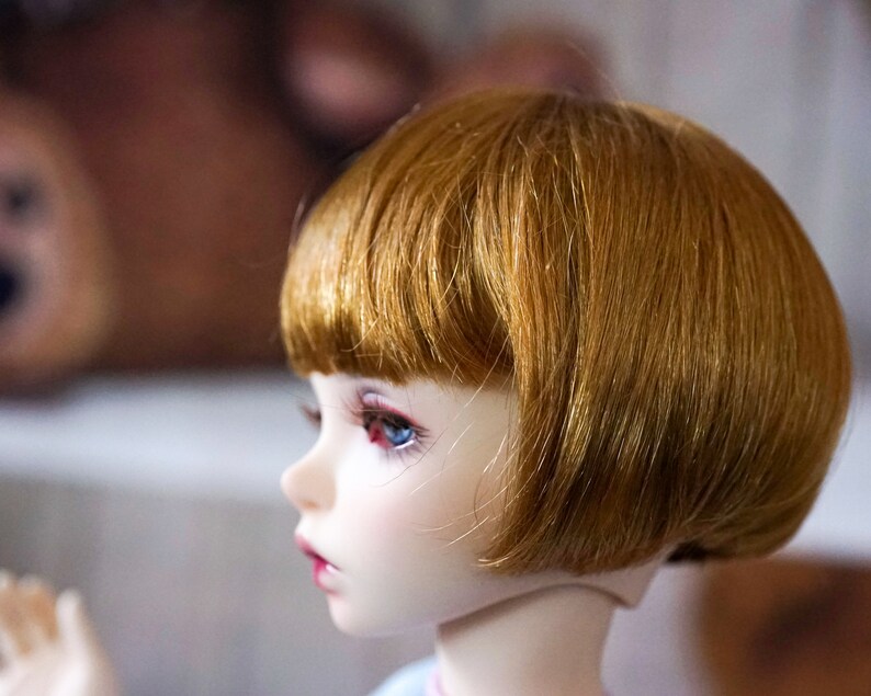 May include: A doll with short, straight, auburn hair styled in a bob. The doll has blue eyes and a light skin tone.