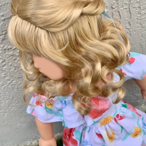 May include: A doll with long, wavy, blonde hair styled in an updo with a light blue ribbon. The doll is wearing a white dress with a floral pattern in red, blue, and yellow. The dress has an off-the-shoulder neckline.