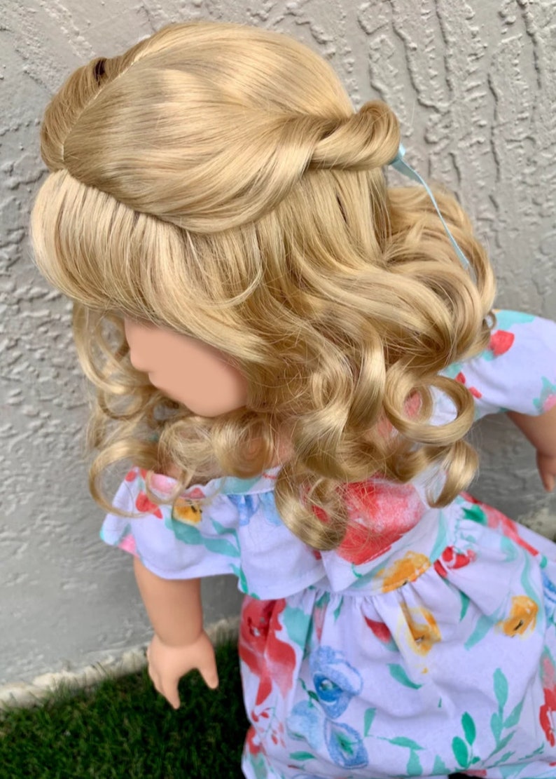 May include: A doll with long, wavy, blonde hair styled in an updo with a light blue ribbon. The doll is wearing a white dress with a floral pattern in red, blue, and yellow. The dress has an off-the-shoulder neckline.