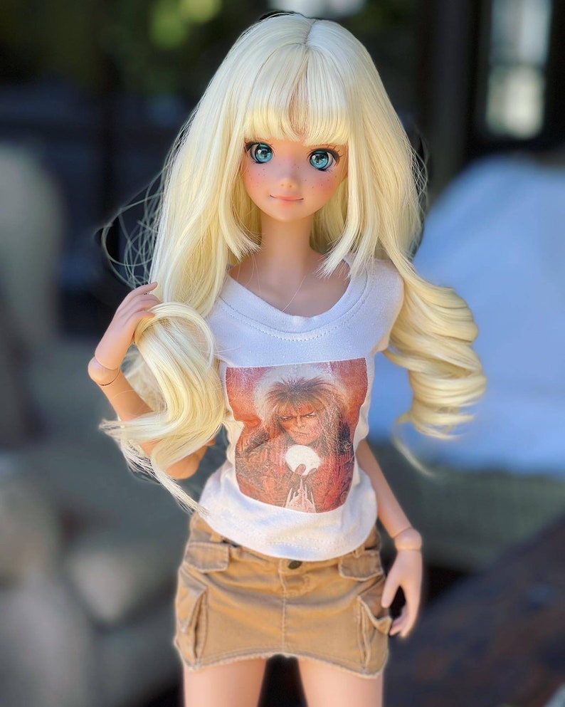 May include: A blonde doll wearing a white t-shirt with a printed image of a man with long hair and a beard. The doll is also wearing a brown skirt and has blue eyes.