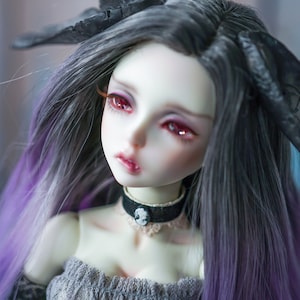 May include: A doll with long, dark gray and purple hair, wearing a black choker with a skull charm. The doll has red eyes and a delicate, porcelain-like face.