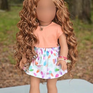 K&ouml;nnte beinhalten: Eine Puppe mit langem, lockigem, hellbraunem Haar tr&auml;gt ein pfirsichfarbenes Kurzarm-Oberteil und einen wei&szlig;en Rock mit einem bunten, abstrakten Muster aus kleinen, abgerundeten Formen. Die Puppe tr&auml;gt eine silberfarbene Kette, Perlenarmb&auml;nder und goldene Sandalen.