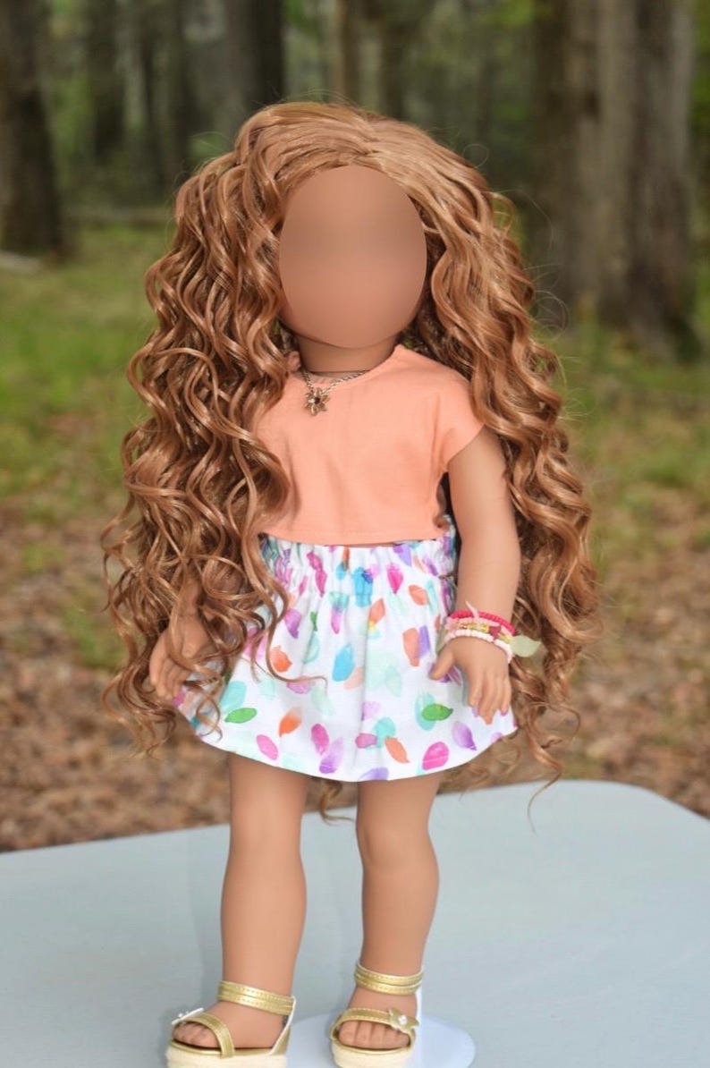 K&ouml;nnte beinhalten: Eine Puppe mit langem, lockigem, hellbraunem Haar tr&auml;gt ein pfirsichfarbenes Kurzarm-Oberteil und einen wei&szlig;en Rock mit einem bunten, abstrakten Muster aus kleinen, abgerundeten Formen. Die Puppe tr&auml;gt eine silberfarbene Kette, Perlenarmb&auml;nder und goldene Sandalen.