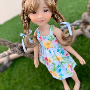 May include: A doll with long brown hair in braids, wearing a white dress with a floral pattern. The doll is smiling and looking at the camera.