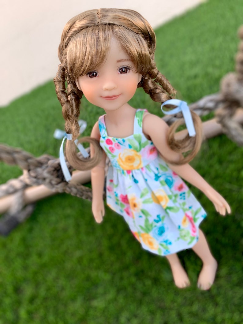 May include: A doll with long brown hair in braids, wearing a white dress with a floral pattern. The doll is smiling and looking at the camera.