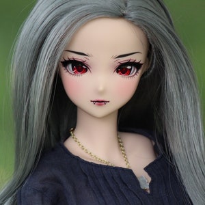 May include: A doll with long, silver-grey hair, wearing a black blouse and a gold chain necklace. The doll has red eyes and a painted-on smile with fangs.