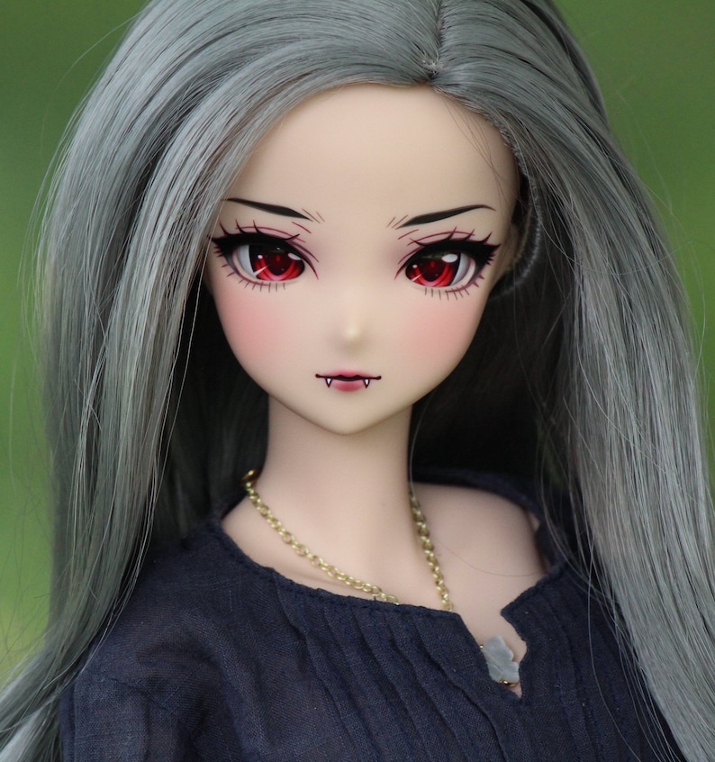 May include: A doll with long, silver-grey hair, wearing a black blouse and a gold chain necklace. The doll has red eyes and a painted-on smile with fangs.
