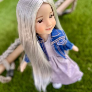 May include: A doll with long, silver hair, wearing a light purple dress and a blue denim jacket. The doll has brown eyes and is positioned on a swing, with a green background.