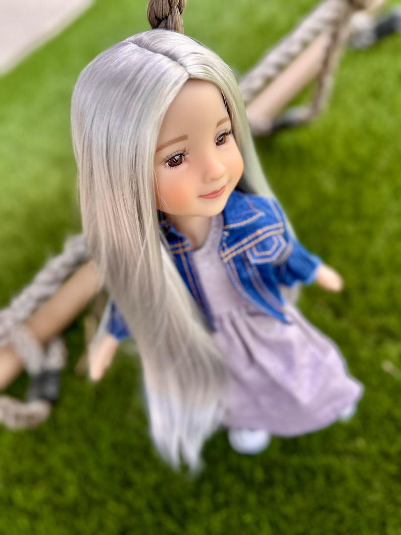 May include: A doll with long, silver hair, wearing a light purple dress and a blue denim jacket. The doll has brown eyes and is positioned on a swing, with a green background.
