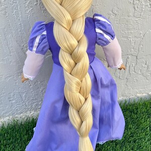 May include: A doll wearing a purple dress with a long, blonde braid. The dress has white trim and puffy sleeves. The doll is standing on green grass.