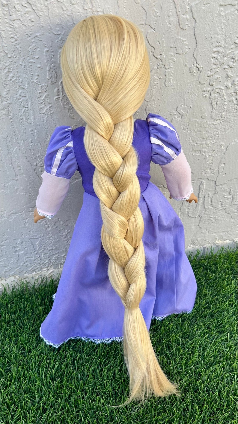 May include: A doll wearing a purple dress with a long, blonde braid. The dress has white trim and puffy sleeves. The doll is standing on green grass.