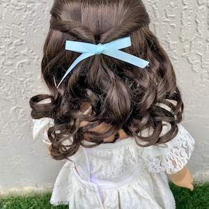 May include: A doll with long brown curly hair styled with a light blue bow. The doll is wearing a white lace dress.