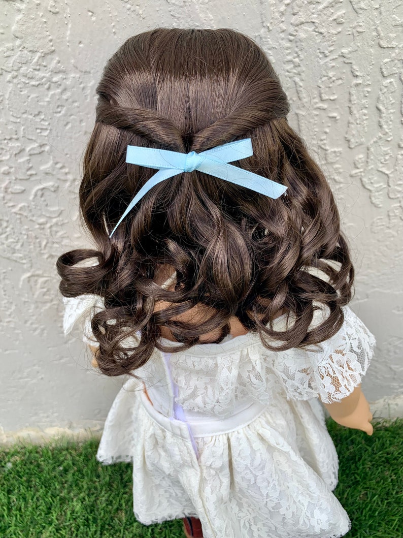 May include: A doll with long brown curly hair styled with a light blue bow. The doll is wearing a white lace dress.