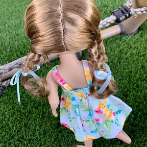 May include: A doll with long brown hair styled in two braids with light blue ribbons. The doll is wearing a white dress with a floral pattern in shades of yellow, pink, and green.