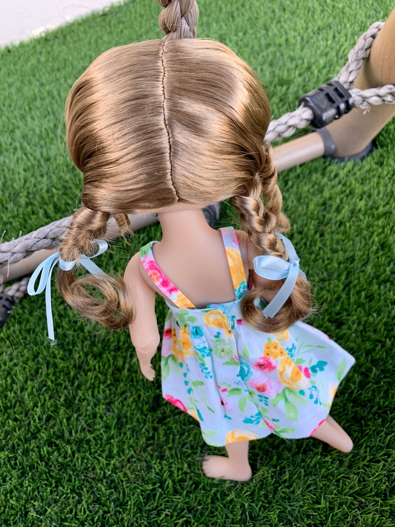 May include: A doll with long brown hair styled in two braids with light blue ribbons. The doll is wearing a white dress with a floral pattern in shades of yellow, pink, and green.