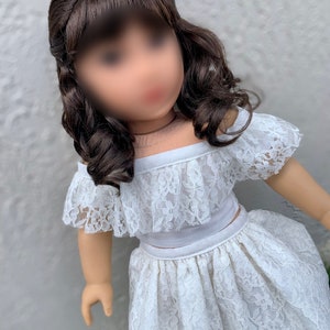 May include: A white lace off-the-shoulder top and skirt set for a doll. The top has a ruffled neckline and the skirt is gathered at the waist.