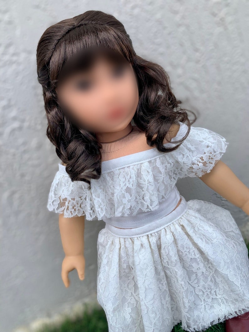 May include: A white lace off-the-shoulder top and skirt set for a doll. The top has a ruffled neckline and the skirt is gathered at the waist.