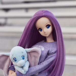 May include: A doll with purple hair wearing a lavender sweatshirt with a white graphic and holding a blue plush elephant toy.