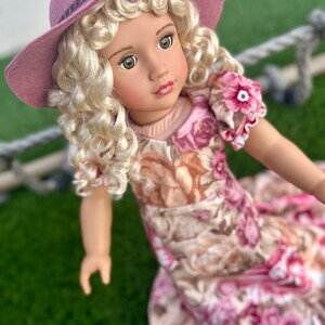 May include: A doll with blonde curly hair wearing a pink floral dress and a wide-brimmed pink hat.