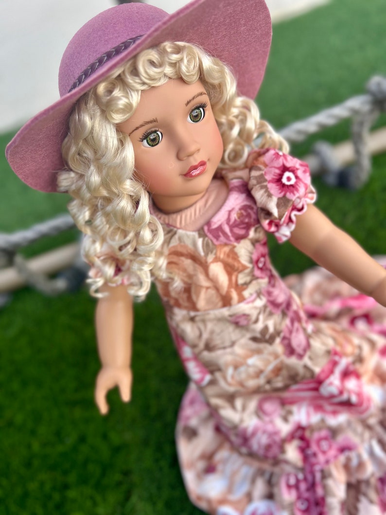May include: A doll with blonde curly hair wearing a pink floral dress and a wide-brimmed pink hat.