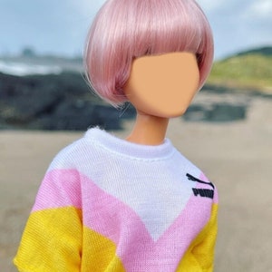 Peut inclure: Une poup&eacute;e avec des cheveux roses coiff&eacute;s en coupe au carr&eacute; portant un pull ray&eacute; jaune, rose et blanc.