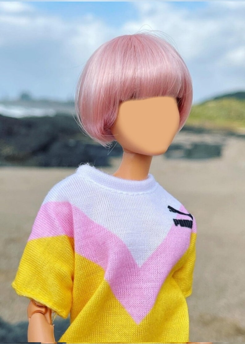 Peut inclure: Une poup&eacute;e avec des cheveux roses coiff&eacute;s en coupe au carr&eacute; portant un pull ray&eacute; jaune, rose et blanc.