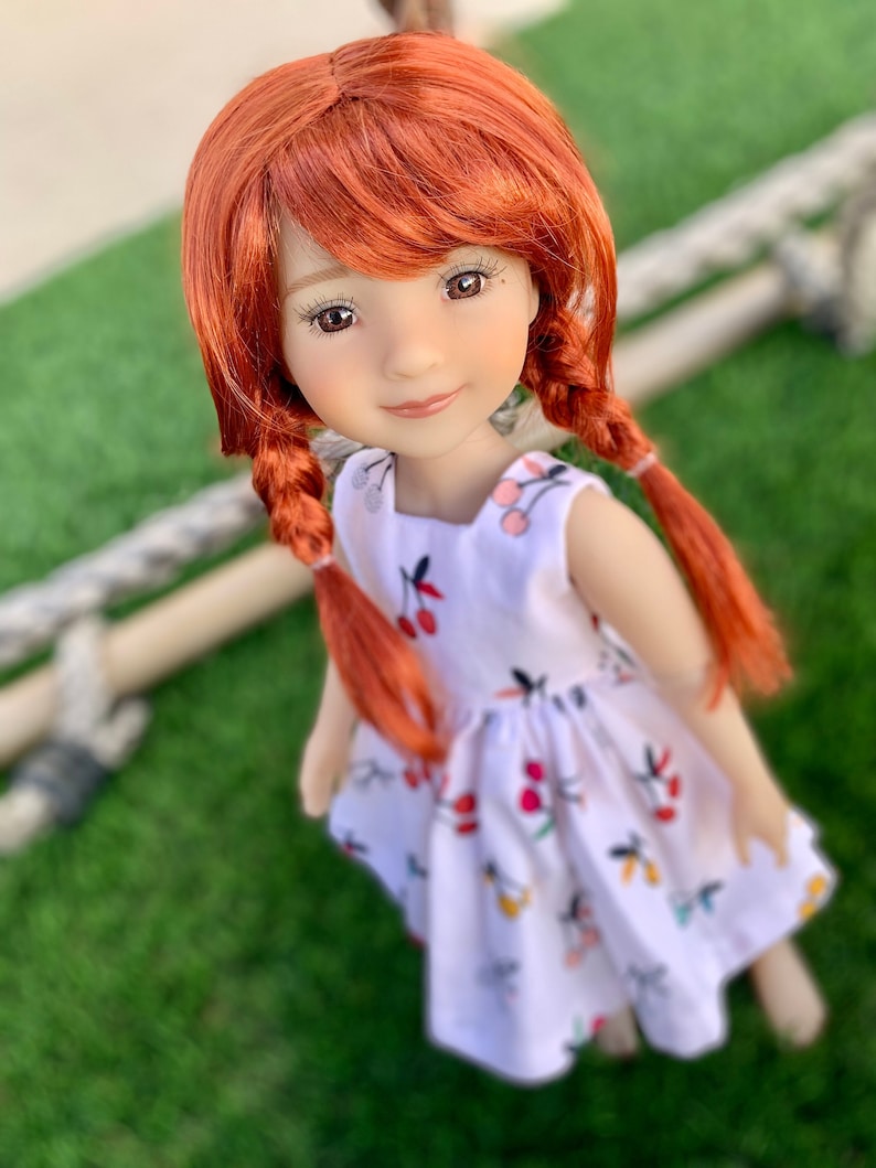 May include: A doll with long, bright red hair styled in braids, wearing a white dress with a cherry print. The doll has brown eyes and a sweet smile.