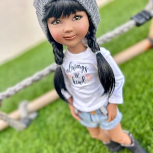 May include: A doll with long black hair in braids wearing a gray beanie, a white t-shirt with the text "Always Be Kind" and denim shorts. The doll is wearing black boots and is standing on green grass.