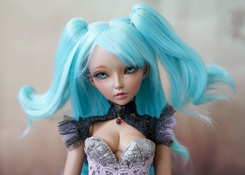 Puede incluir: Una mu&ntilde;eca con el pelo largo y azul brillante peinado en coletas. La mu&ntilde;eca lleva un vestido negro y plateado con un corpi&ntilde;o de encaje y un collar oscuro con una piedra preciosa p&uacute;rpura.