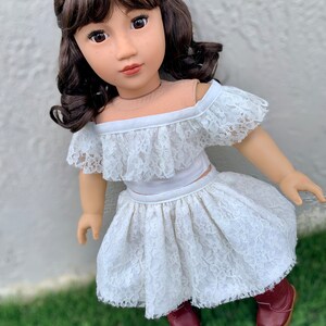May include: A doll wearing a white lace off-the-shoulder top and skirt. The doll has brown hair and brown eyes.
