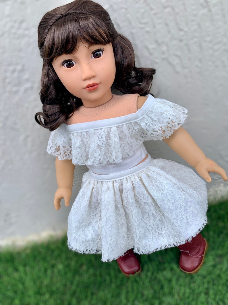 May include: A doll wearing a white lace off-the-shoulder top and skirt. The doll has brown hair and brown eyes.