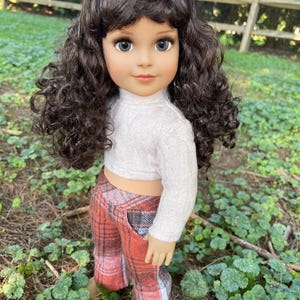 May include: A doll with brown curly hair, wearing a white sweater and plaid pants in shades of red, orange, and gray. The doll is standing outdoors in a grassy area.