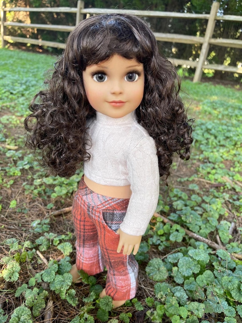 May include: A doll with brown curly hair, wearing a white sweater and plaid pants in shades of red, orange, and gray. The doll is standing outdoors in a grassy area.