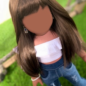 Peut inclure: Une poupée avec de longs cheveux bruns, portant un haut blanc à épaules dénudées, un jean bleu déchiré et un bracelet blanc. La poupée porte des boucles d'oreilles avec des pierres bleues.