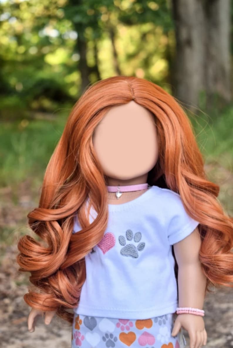 May include: A doll with long, curly, auburn hair wearing a white t-shirt with a pink heart and gray paw print design. The doll is also wearing a gray and orange skirt with paw prints and hearts. The doll is wearing a pink choker with a silver heart charm and a pink beaded bracelet.