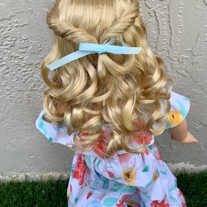 May include: A doll with blonde, curly hair styled with a twisted crown and a light blue ribbon. The doll is wearing a white floral print jumpsuit with red, yellow, and blue flowers.